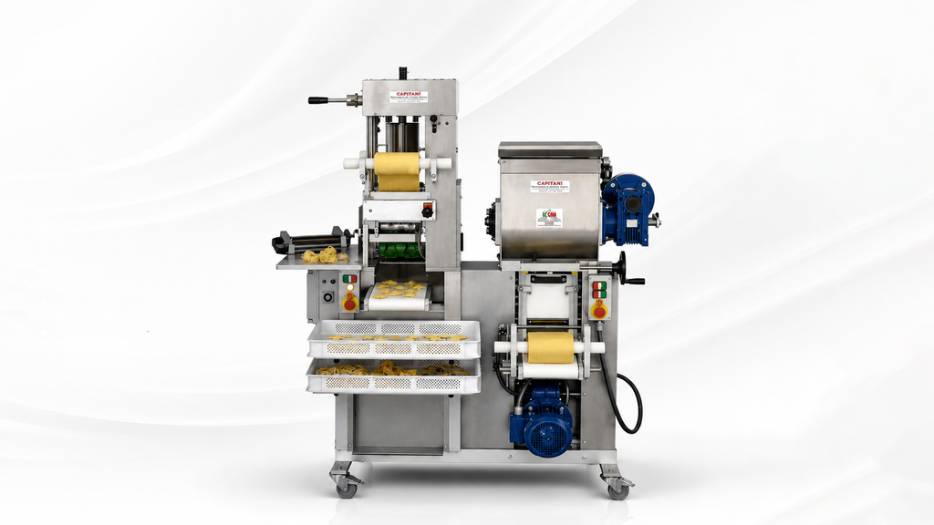 A stainless steel commercial pasta machine sits on a white marble countertop against a white backdrop, surrounded by white trays filled with various types of fresh pasta, including long strands, small shaped pieces, and stuffed ravioli.