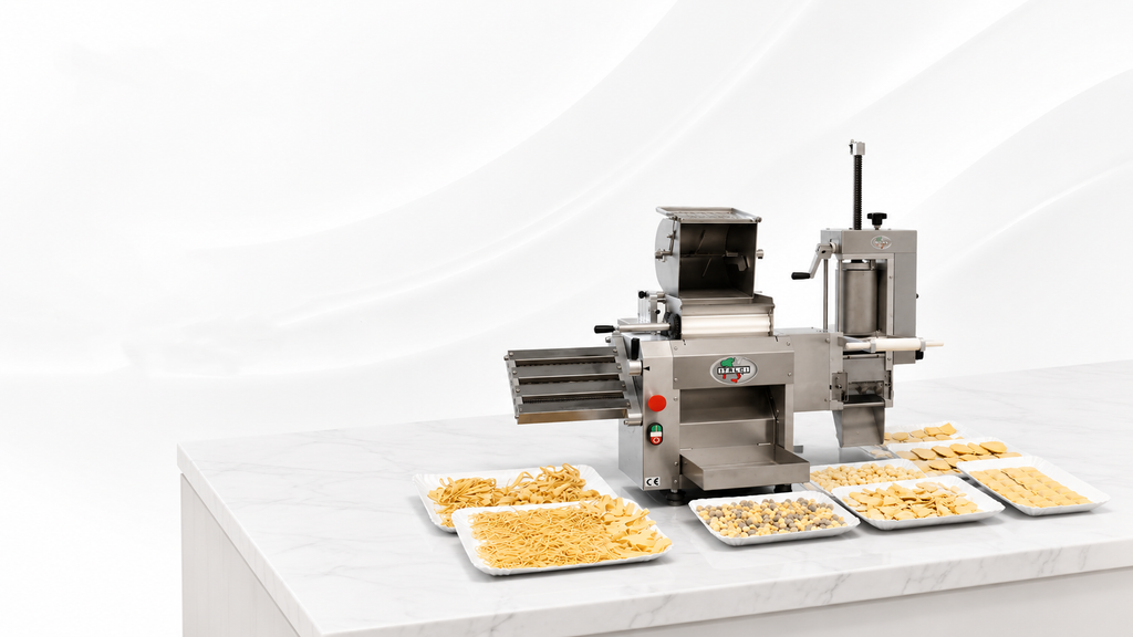 A stainless steel commercial pasta machine sits on a white marble countertop against a white backdrop, surrounded by white trays filled with various types of fresh pasta, including long strands, small shaped pieces, and stuffed ravioli.